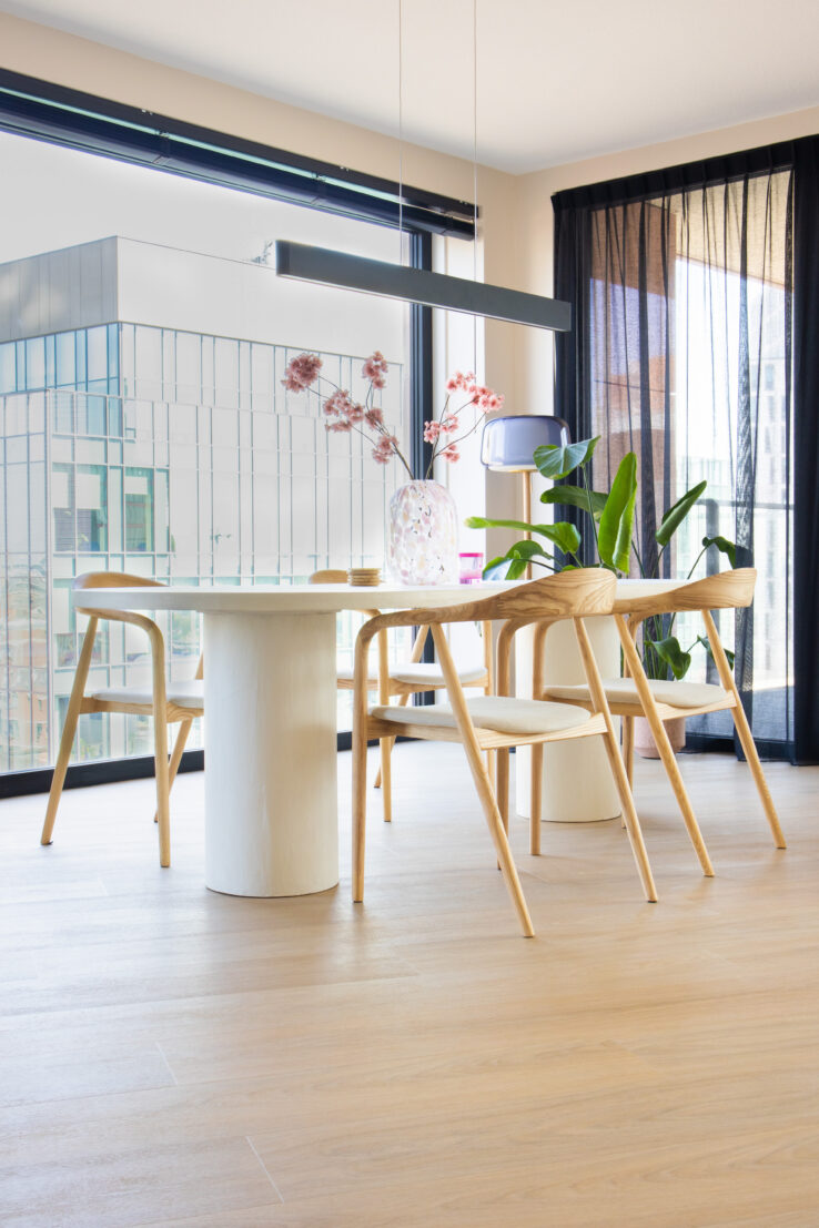 Lichte eiken PVC-vloer in een moderne, zonnige eetkamer met houten stoelen, een ronde witte tafel en grote raamwanden.
