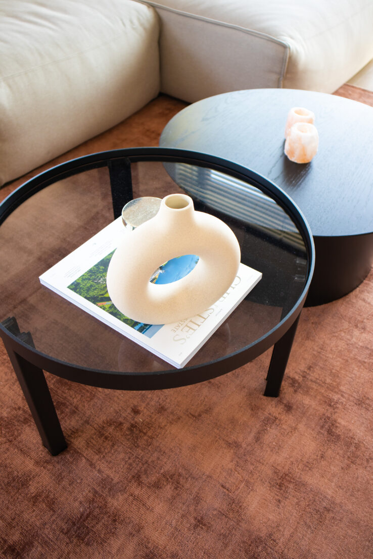 Ronde salontafel met glazen blad, een beige decoratieve vaas en een koffietafelboek op een zachtbruin vloerkleed, naast een zwarte bijzettafel.