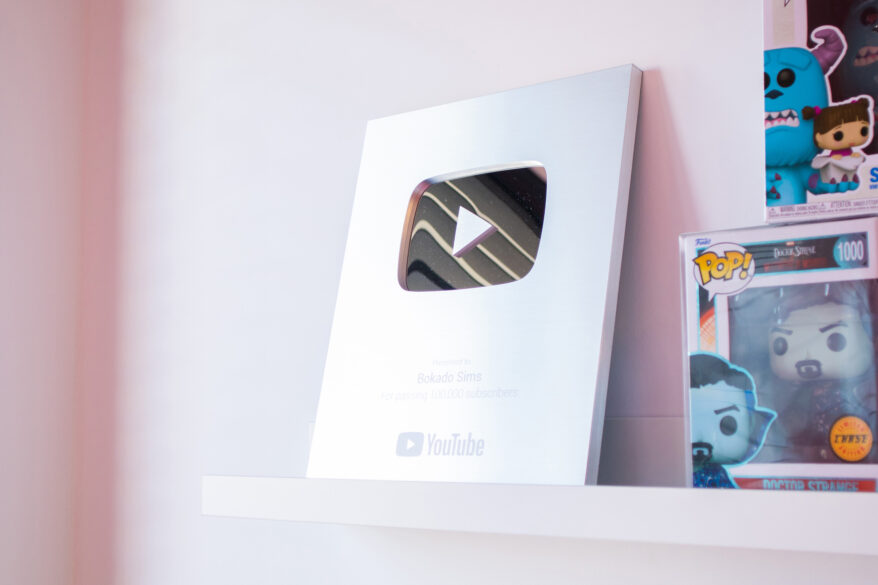 YouTube Silver Play Button op een witte plank, naast Funko Pop-figuren in een kleurrijke kast.