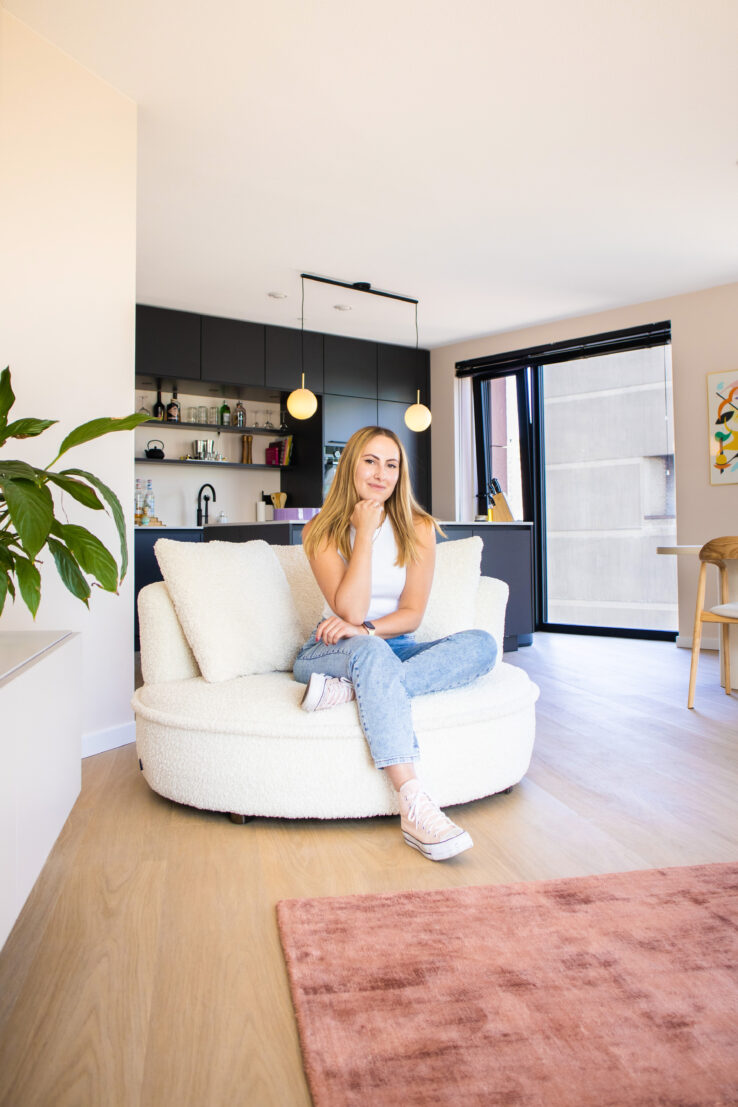 Vrouw zittend op een ronde witte fauteuil in een moderne woonkamer met lichte eiken PVC-vloer en zwarte keuken op de achtergrond.