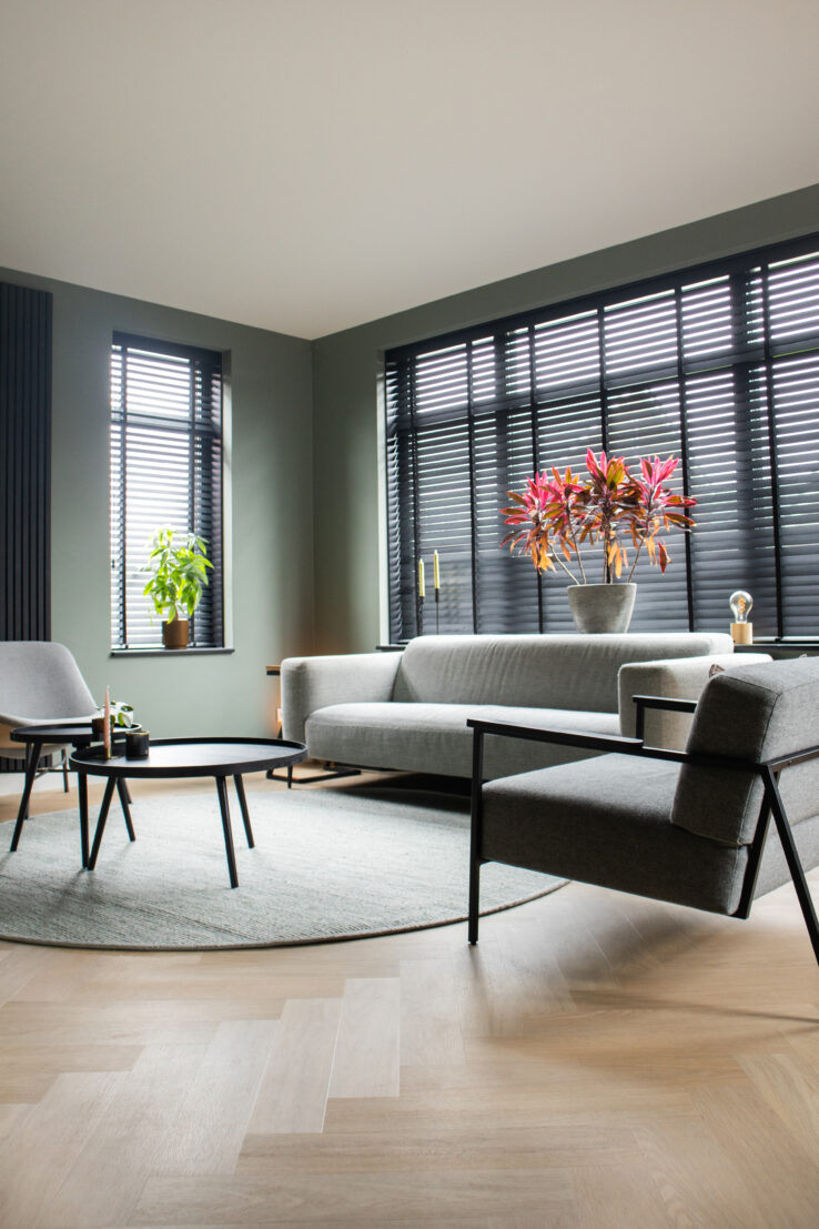 Moderne woonkamer met een lichte eiken visgraat PVC-vloer, grijze zitmeubels, zwarte jaloezieën en een ronde salontafel met planten.