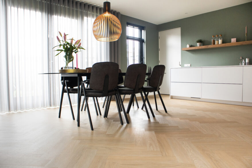 Lichte eiken visgraat PVC-vloer in een moderne eetkamer met zwarte stoelen, een houten hanglamp en witte kastenwand met groene muur.