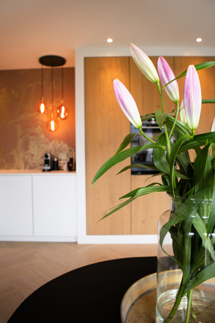 Close-up van roze lelies in een glazen vaas, met op de achtergrond een moderne keuken met houten kasten en warme verlichting.