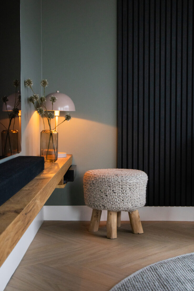 Hoek met lichte eiken visgraat PVC-vloer, houten wandplank met lamp en vaas, en een ronde gevlochten kruk op houten pootjes.
