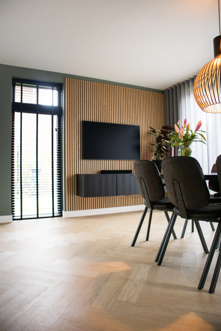 Lichte eiken visgraat PVC-vloer in een moderne woonkamer met een houten lamellenwand, tv-meubel en donkere eetkamerstoelen naast een grote vaas met bloemen.