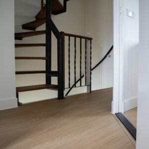 Open trap met houten treden en zwart metalen leuning, naast een vloer bedekt met lichte PVC-vloer. De ruimte heeft een moderne uitstraling met neutrale kleuren en eenvoudige afwerking.