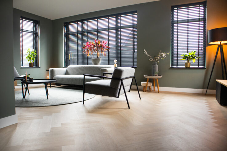 Lichte eiken visgraatvloer in een stijlvolle woonkamer met donkergroene muren, zwarte jaloezieën en modern grijze meubels.