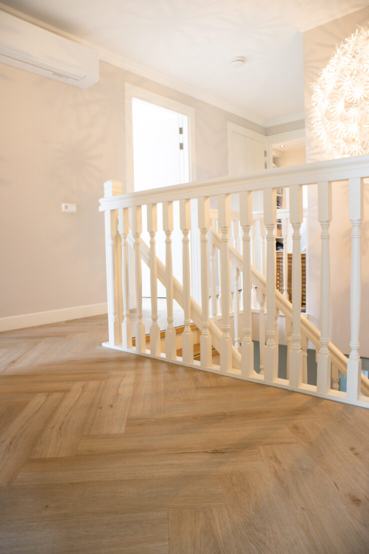 Lichte eiken visgraat PVC-vloer op een overloop met witte trapleuning en zachtbeige wanden.