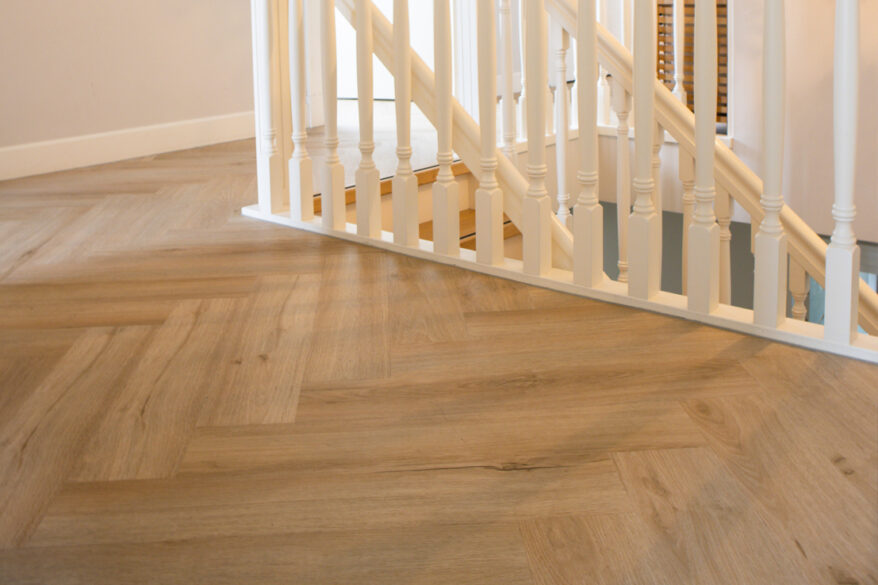 Close-up van een lichte eiken visgraat PVC-vloer naast een witte trapleuning op de overloop.