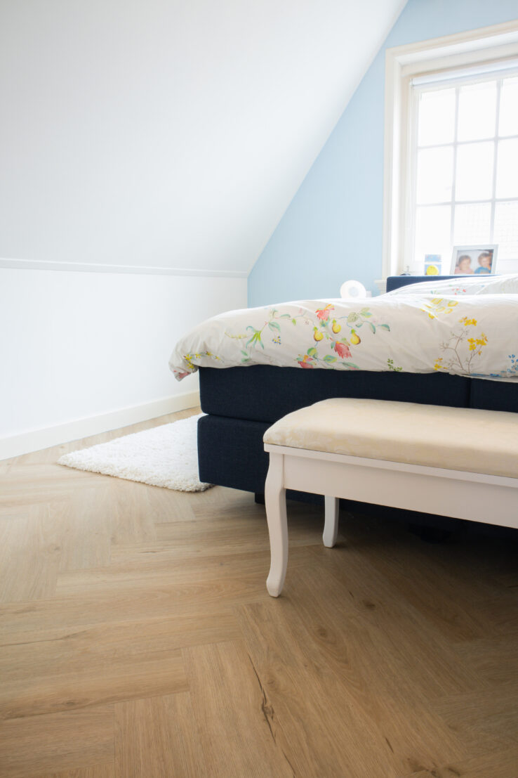 Slaapkamer met lichte eiken visgraat PVC-vloer, een bed met bloemenbeddengoed en een wit bankje aan het voeteneind.