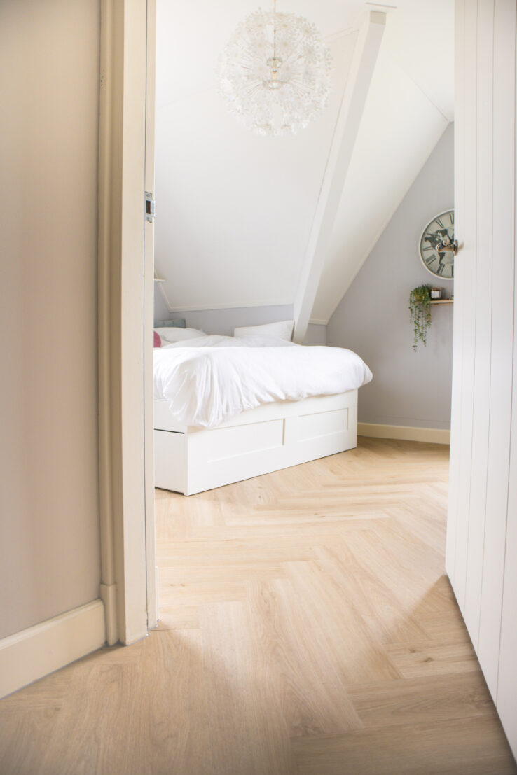 Slaapkamer met lichte eiken visgraat PVC-vloer, een wit bed en schuine plafonds in zachte grijstinten.