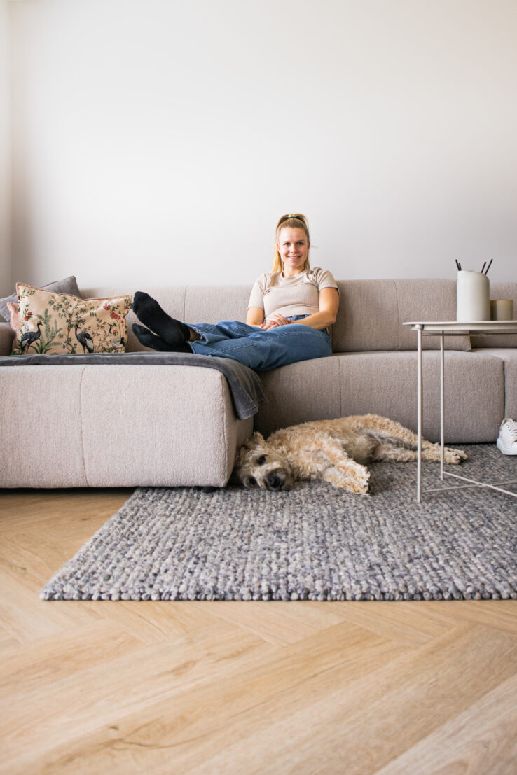 Vrouw ontspannen op een beige hoekbank met een kleed op de lichte visgraatvloer, terwijl haar hond rustig op het vloerkleed ligt te slapen.