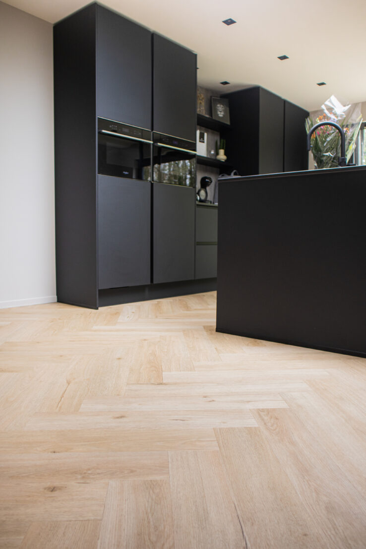 Lichte eiken visgraat PVC-vloer in een moderne keuken met matzwarte kasten en kookeiland.