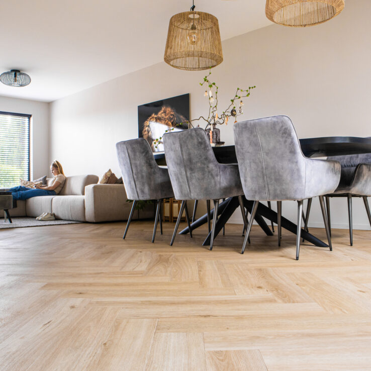 Lichte eiken visgraat PVC-vloer in een ruime woonkamer met eetkamerstoelen, zwarte eettafel en een zithoek met bank op de achtergrond.