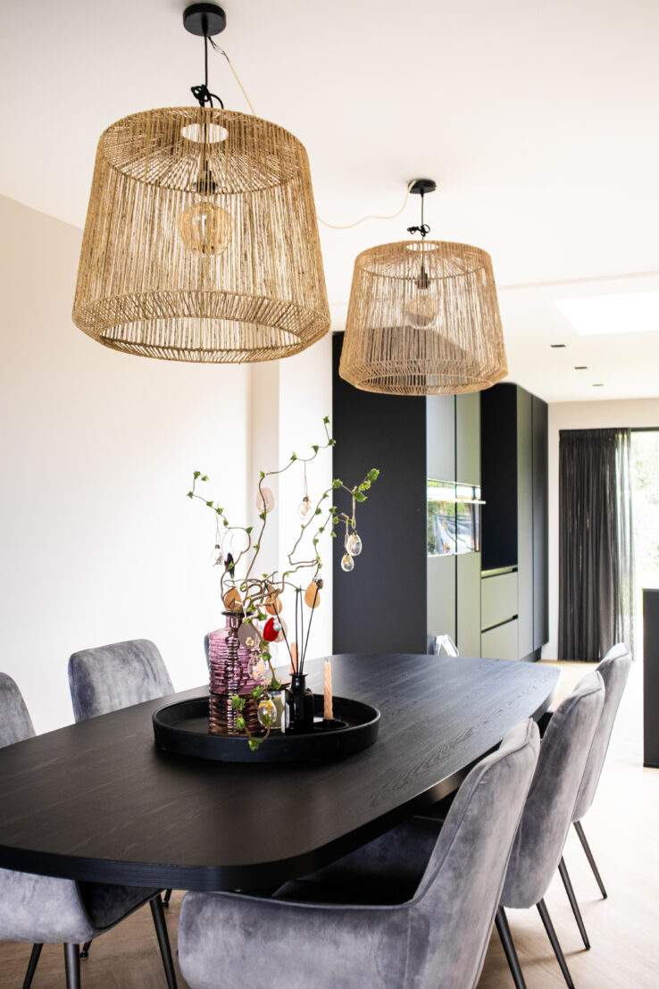 Eetkamer met een zwarte ovale tafel, grijze fluwelen stoelen en twee grote hanglampen van natuurlijk touw, met op de tafel een decoratief dienblad met vazen en takken.