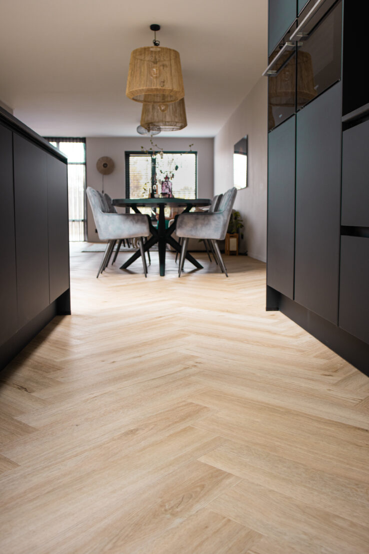 Lichte eiken visgraat PVC-vloer tussen twee zwarte keukenblokken, met zicht op de eethoek met grijze stoelen en rieten hanglampen.