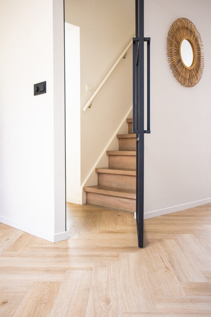 Hal met lichte visgraatvloer, zwart stalen deur en houten trap met witte leuning voor een frisse, moderne uitstraling.