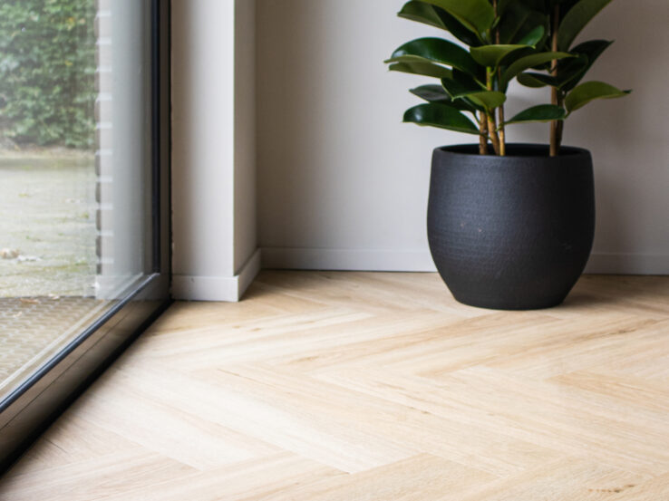 Lichte eiken PVC-vloer met een plantenpot in de hoek, naast een grote schuifdeur die natuurlijk licht binnenlaat.