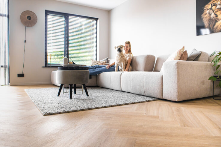 Lichte eiken visgraat PVC-vloer in een woonkamer met beige hoekbank, vloerkleed en een vrouw met haar hond op de bank.