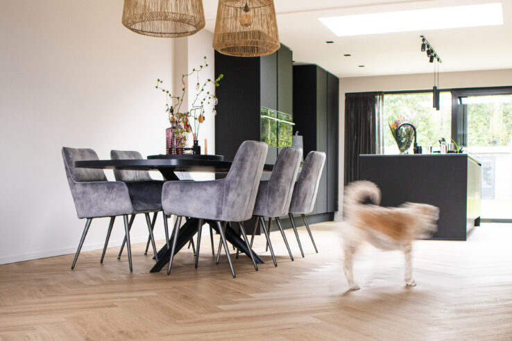 Lichte eiken visgraat PVC-vloer in een moderne woonkeuken met grijze eetkamerstoelen, zwarte tafel en een hond die door de ruimte loopt.