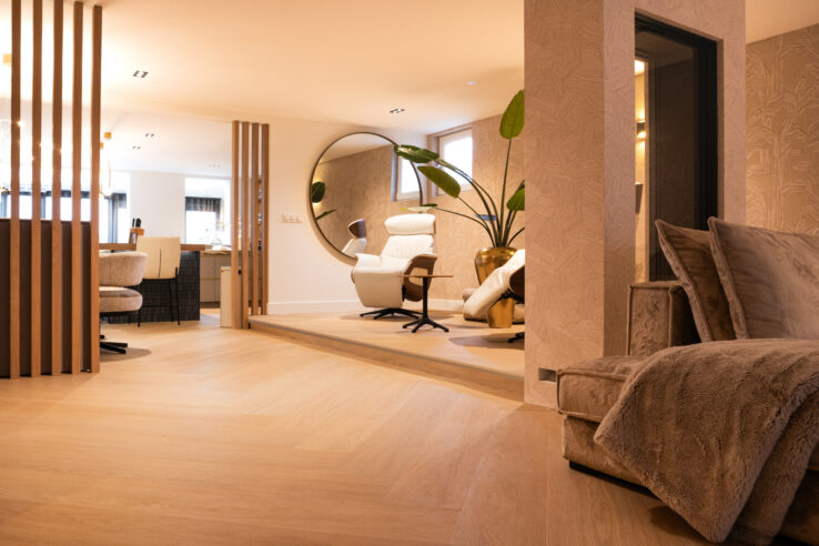 Stijlvolle woonkamer met lichte visgraatvloer, luxe loungestoel voor een grote ronde spiegel en warme beige tinten.