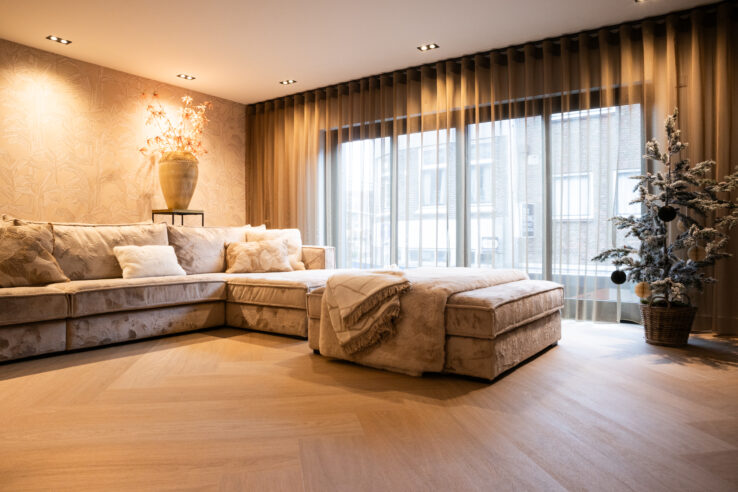 Lichte woonkamer met grote hoekbank in zachte stof, vloer in warm visgraatpatroon en brede raampartij met transparante gordijnen.