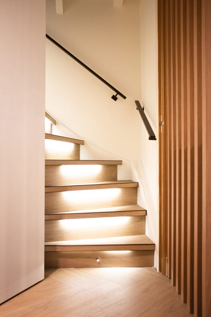 Houten trap met geïntegreerde LED-verlichting onder de treden en een strakke zwarte leuning langs de wand.
