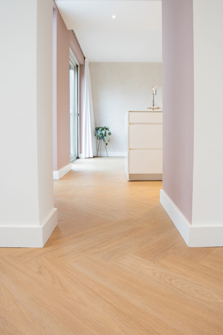 Lichte visgraat PVC-vloer in een moderne doorgang met roze wanden, witte plinten en zicht op een lichte keukenruimte met groot raam en planten.