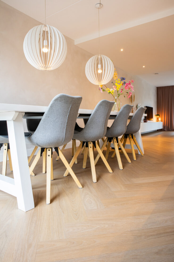 Lichte visgraat PVC-vloer in een moderne eethoek met een lange witte tafel, grijze eetstoelen met houten poten en twee grote ronde hanglampen.