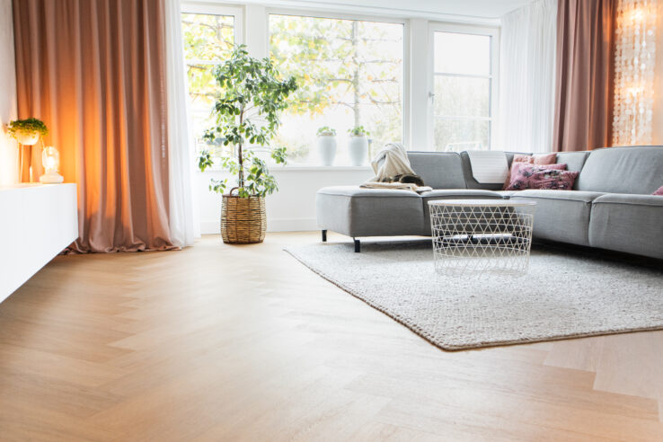 Lichte eiken visgraatvloer in een zachte, moderne woonkamer met een grijze hoekbank, roze gordijnen en veel daglicht.