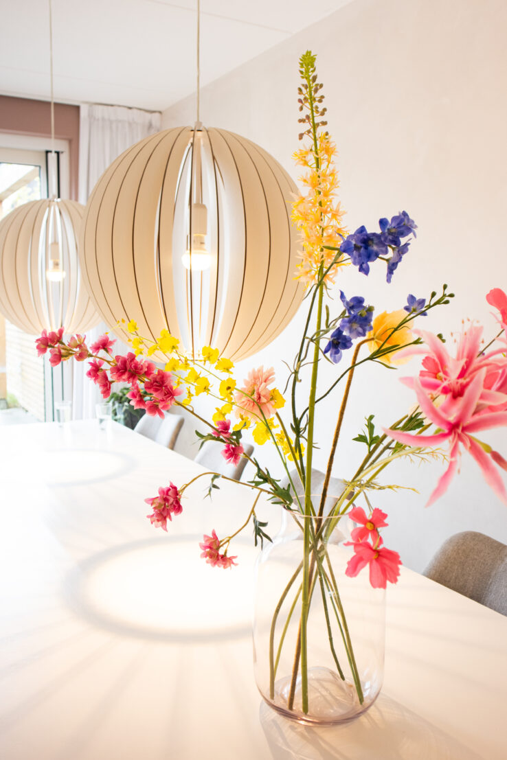 Close-up van een vaas met kleurrijke bloemen op een witte eettafel, met twee grote ronde hanglampen die warm licht verspreiden