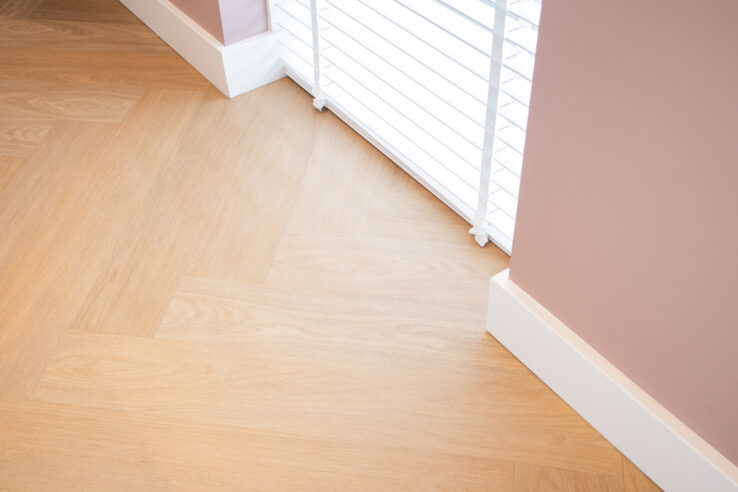 Detail van een lichte visgraat PVC-vloer bij een raam met witte jaloezieën en roze wanden met witte plinten.