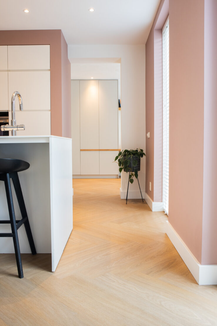Lichte visgraat PVC-vloer in een moderne keuken met roze muren, wit kookeiland, zwarte barkruk en een plant op een metalen standaard richting de hal.