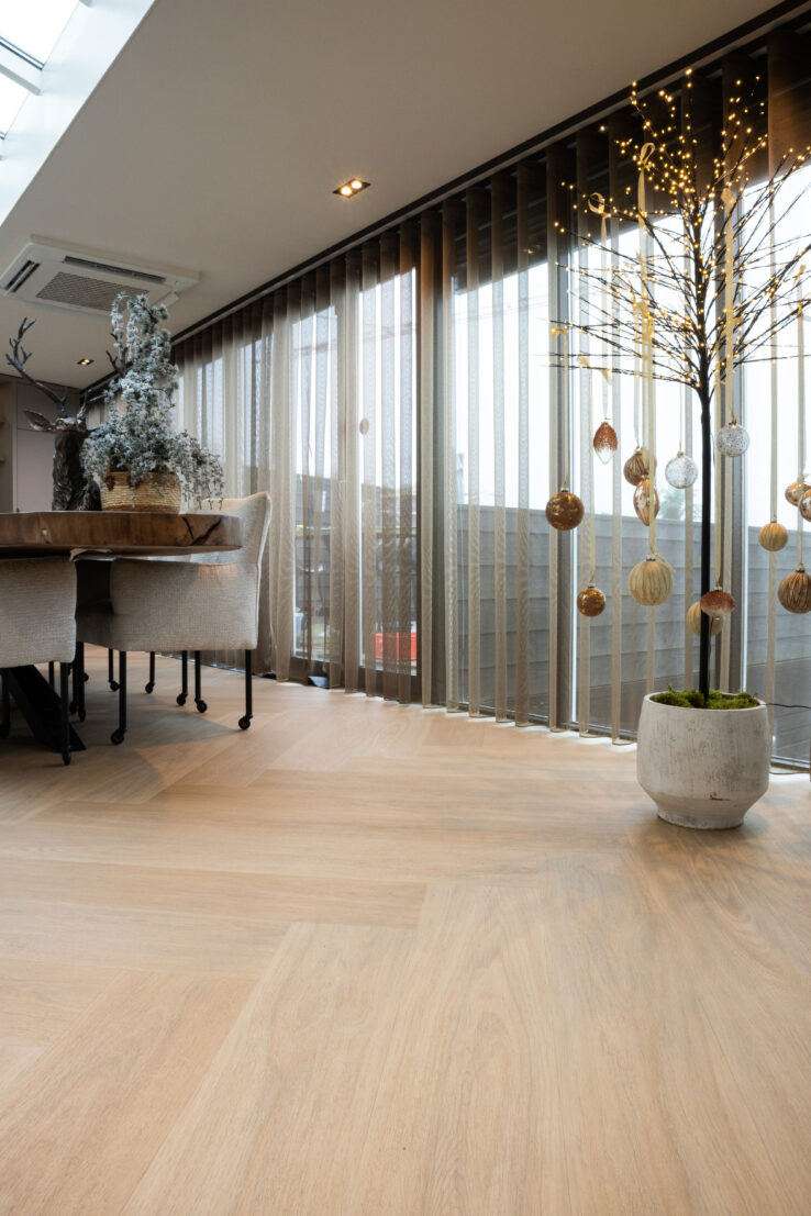 Lichte woonkamer met brede raampartij, warme visgraatvloer en een gedecoreerde boom met hangende ornamenten naast een gezellige eethoek.