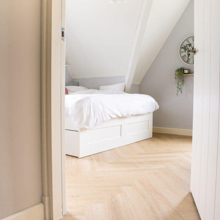 Lichte eiken visgraatvloer in een knusse slaapkamer met schuine wanden, wit bed en zachte grijs-witte tinten.