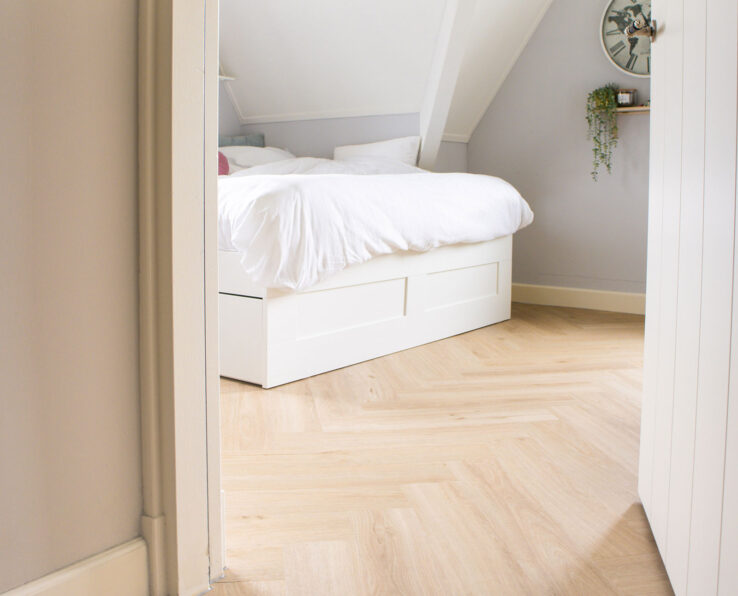 Lichte eiken visgraatvloer in een knusse slaapkamer met schuine wanden, wit bed en zachte grijs-witte tinten.