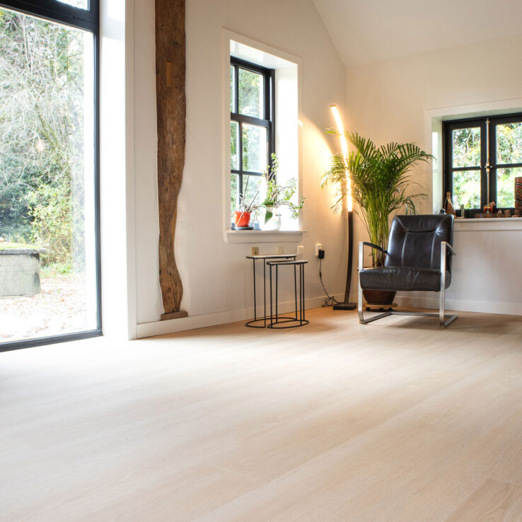 Lichte eiken PVC-vloer in een warm ingerichte woonkamer met zwarte kozijnen, houten balk en een modern leren fauteuil.