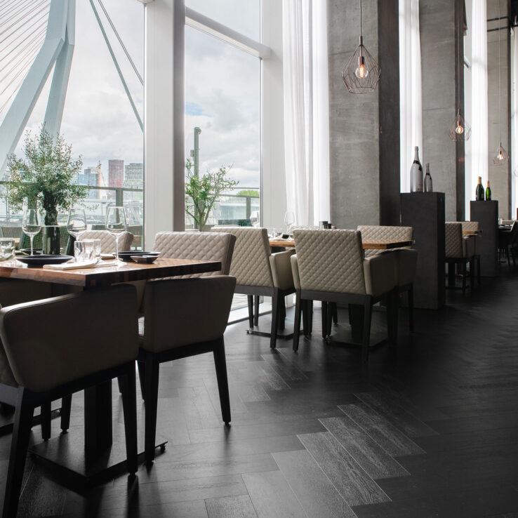 Donkere visgraat PVC-vloer in een modern restaurant met hoge ramen, uitzicht op de Erasmusbrug en stijlvolle, lichte stoelen en houten tafels.