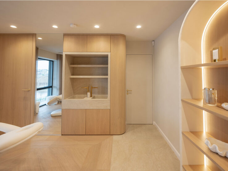 Moderne, minimalistische kamer met houten kasten, een lichte vloer en grote ramen die uitzicht bieden op buiten. De ruimte heeft subtiele verlichting en een open wandplank voor decoratieve objecten.