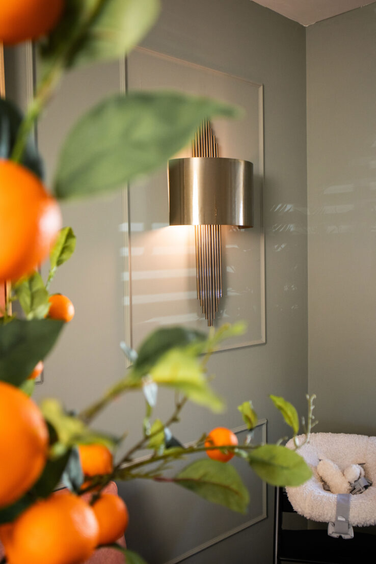 Detail van een moderne wandlamp op een zachtgroene muur, met op de voorgrond takken met oranje vruchten en rechts een babywiegje in beeld.
