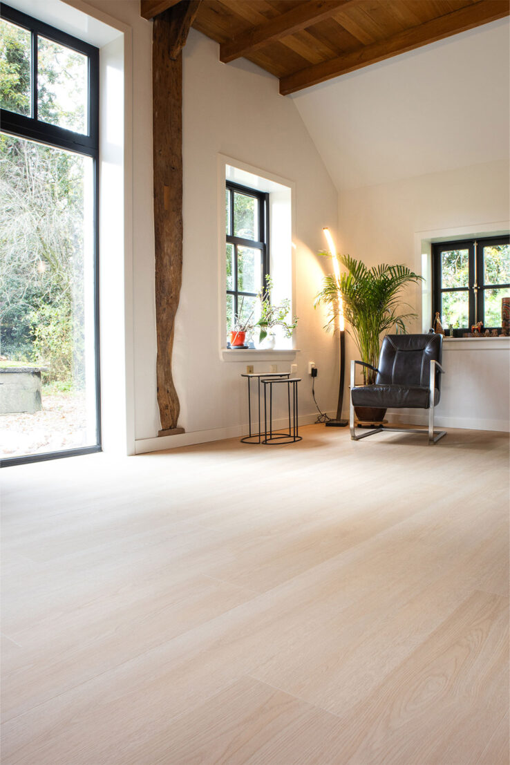 Lichte houtlook PVC-vloer in een ruim, landelijk interieur met grote zwarte ramen, zichtbare houten balken, veel planten en een leren fauteuil in de hoek.