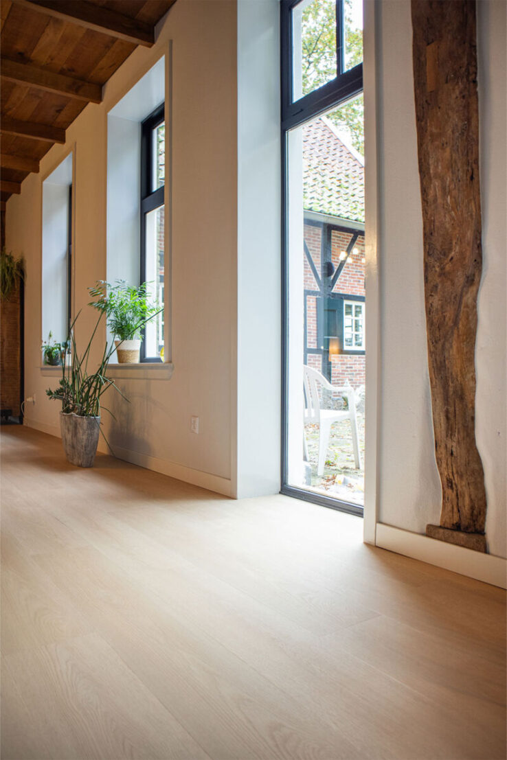 Lichte houtlook PVC-vloer in een lange hal met grote zwarte ramen, houten plafondbalken en diverse planten op de vensterbanken, met uitzicht op een landelijke buitenruimte.