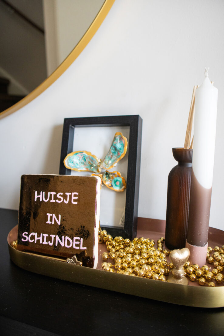 Decoratief plateau met een tegel waarop ‘Huisje in Schijndel’ staat, goudkleurige kralen, kaarsen en een ingelijste kunstprint, voor een ronde spiegel.