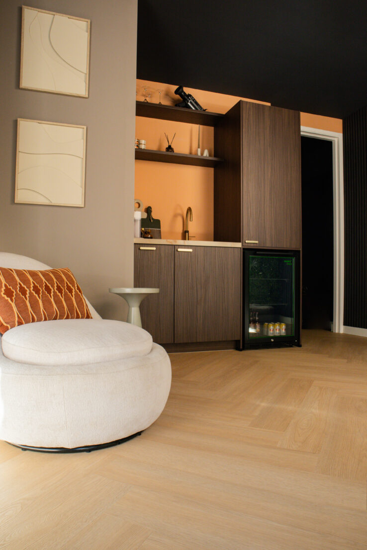 Warme woonkamerhoek met een ronde beige fauteuil, oranje sierkussen en een lichte visgraat PVC-vloer, naast een moderne kitchenette met houtlook kasten en een minibar.