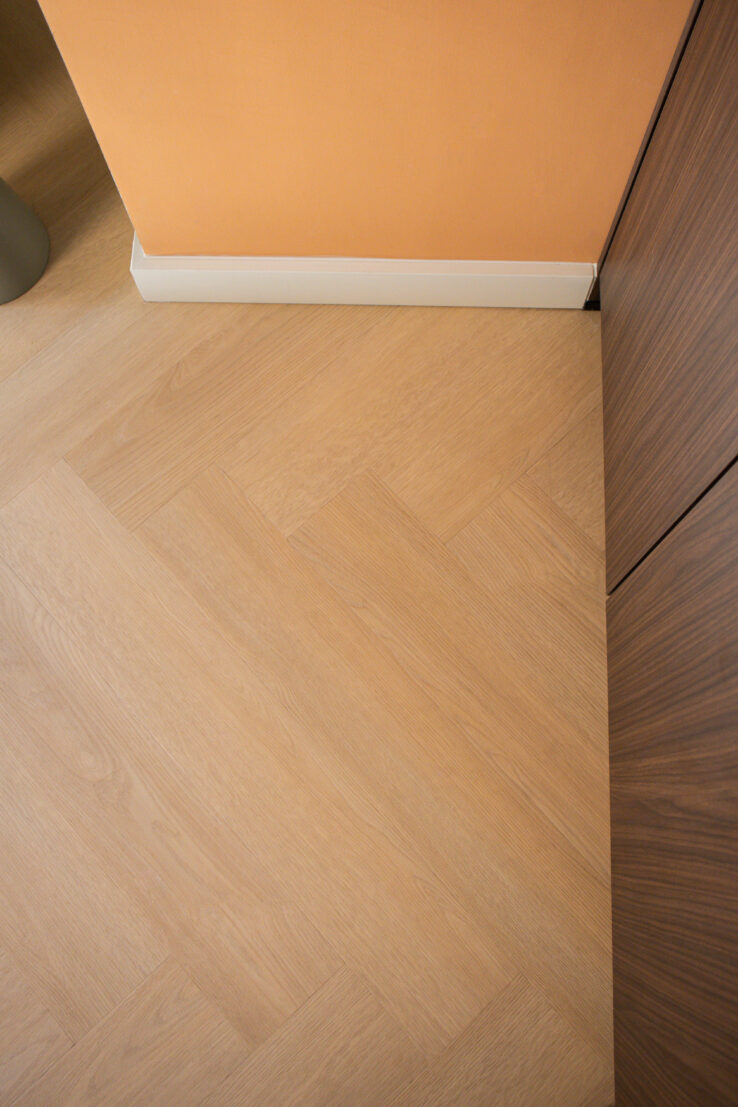 Detail van een lichte visgraat PVC-vloer naast een perzikkleurige muur, witte plint en een houtlook keukenkast.