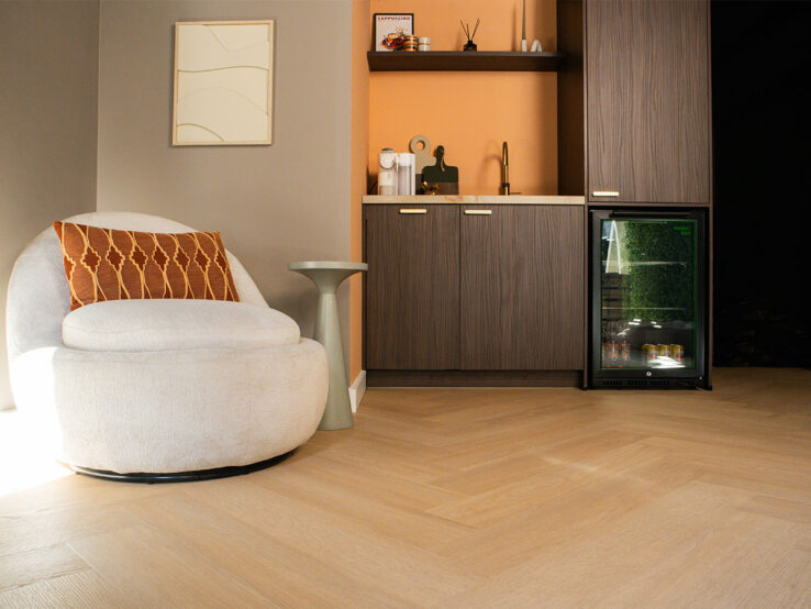 Warme zithoek met een ronde beige fauteuil, oranje sierkussen en een lichte visgraat PVC-vloer, naast een kitchenette met houtlook kasten en een minibar.
