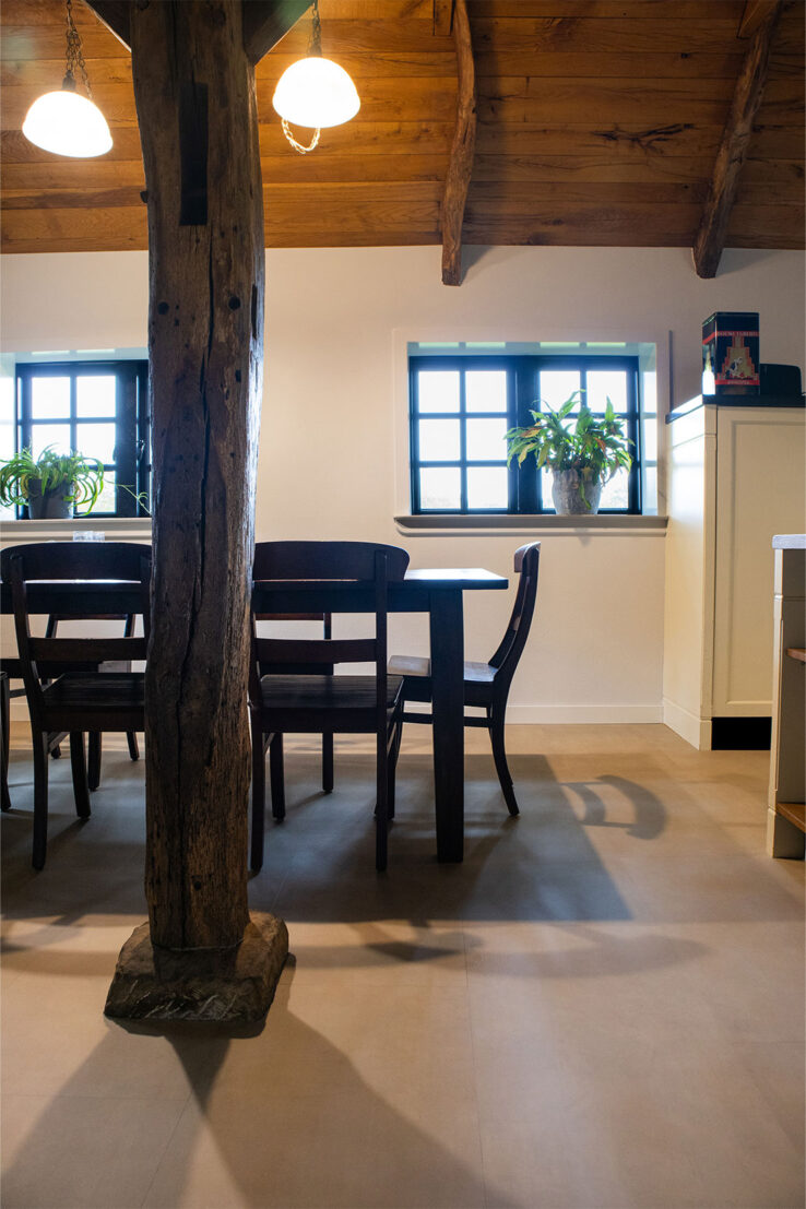 Landelijke eetruimte met houten plafondbalken, een robuuste houten staander, donkere eetkamerstoelen en een rustige beige PVC-vloer, met planten op de vensterbanken.
