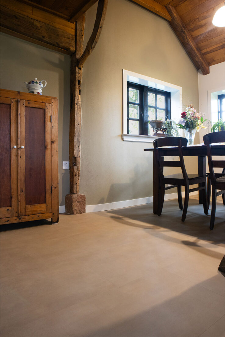 Beige tegel-look PVC-vloer in een landelijke eetkamer met houten plafondbalken, een rustieke houten kast en een donkere eettafel met bloemen op het raamkozijn.