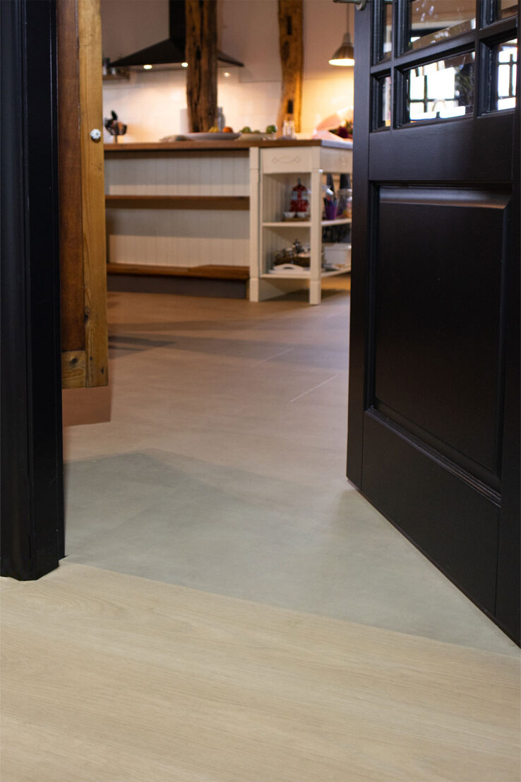 Overgang tussen een lichte houtlook PVC-vloer en een beige tegel-look PVC-vloer bij een zwarte deur, met zicht op een landelijke keuken met houten balken en kookeiland.