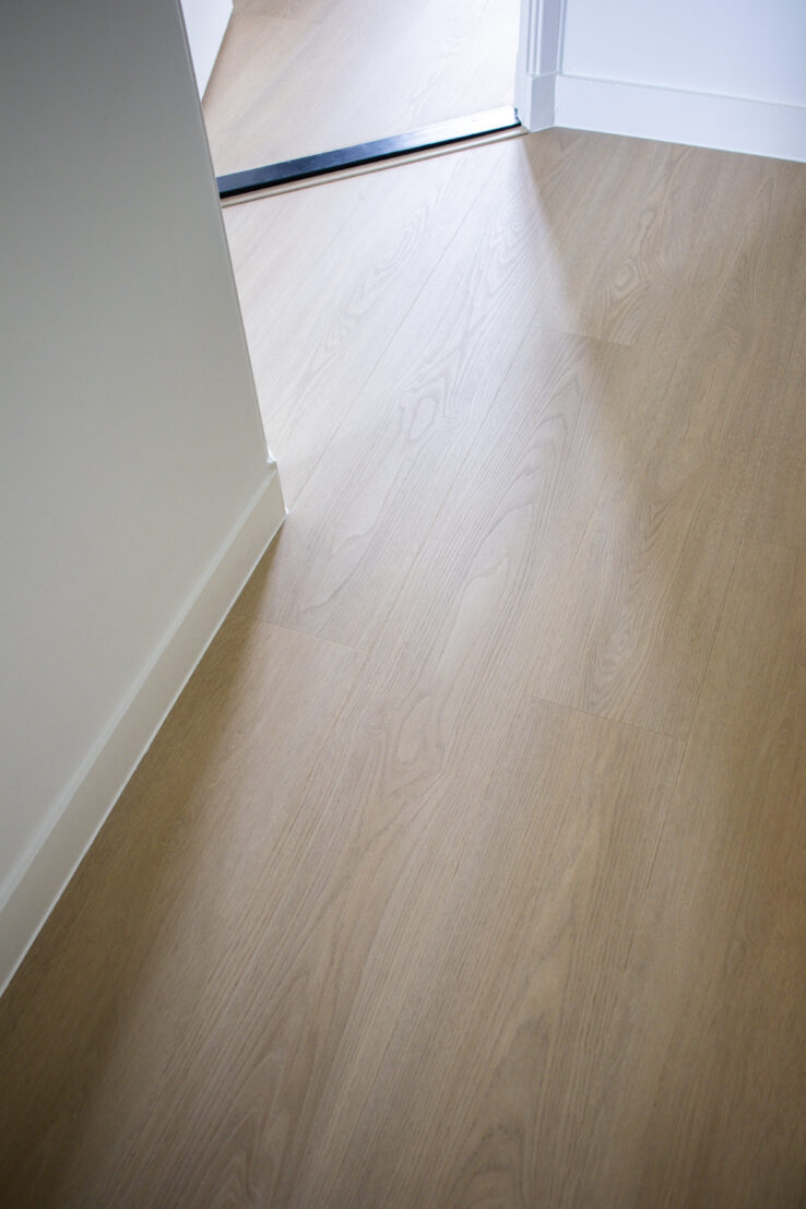 Lichte houtlook PVC-vloer in een doorgang, met subtiele houtnerf en een zwarte overgangsstrip tussen twee ruimtes.
